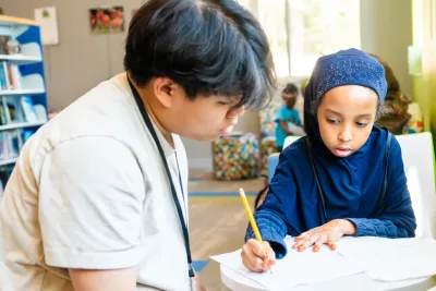 Volunteer tutor watches attentively as young student completes homework in the library